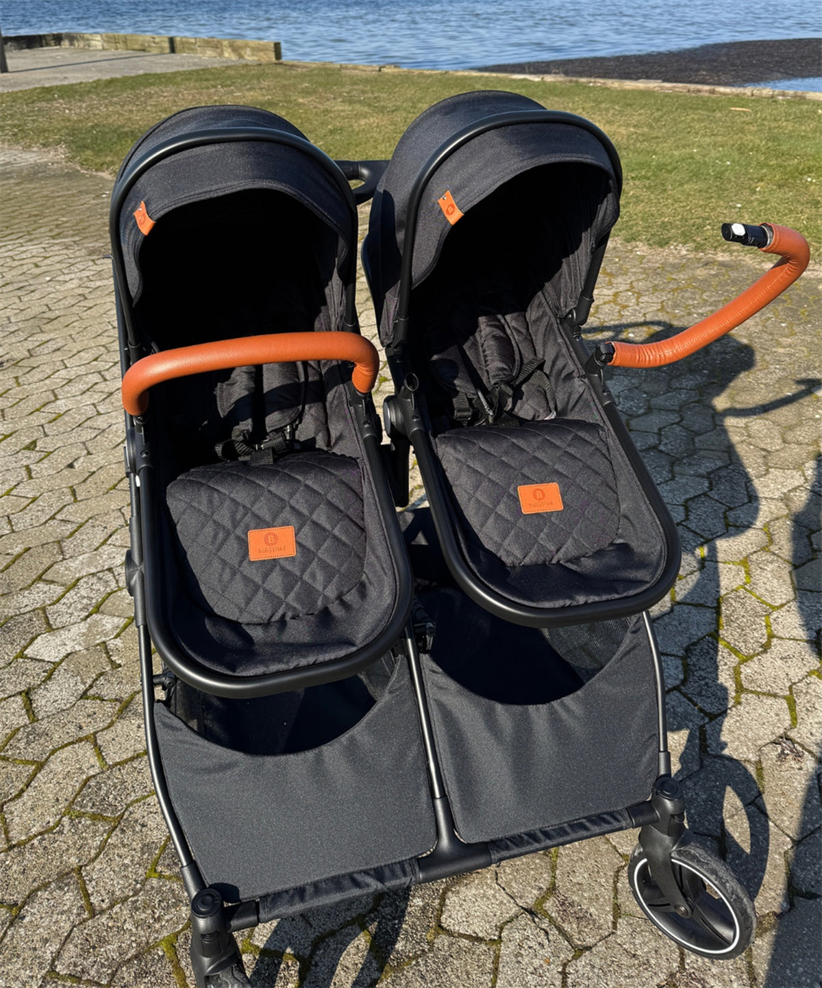 Doppel-Kinderwagen mit schwarzen Sitzen und braunen Griffen am Wasser