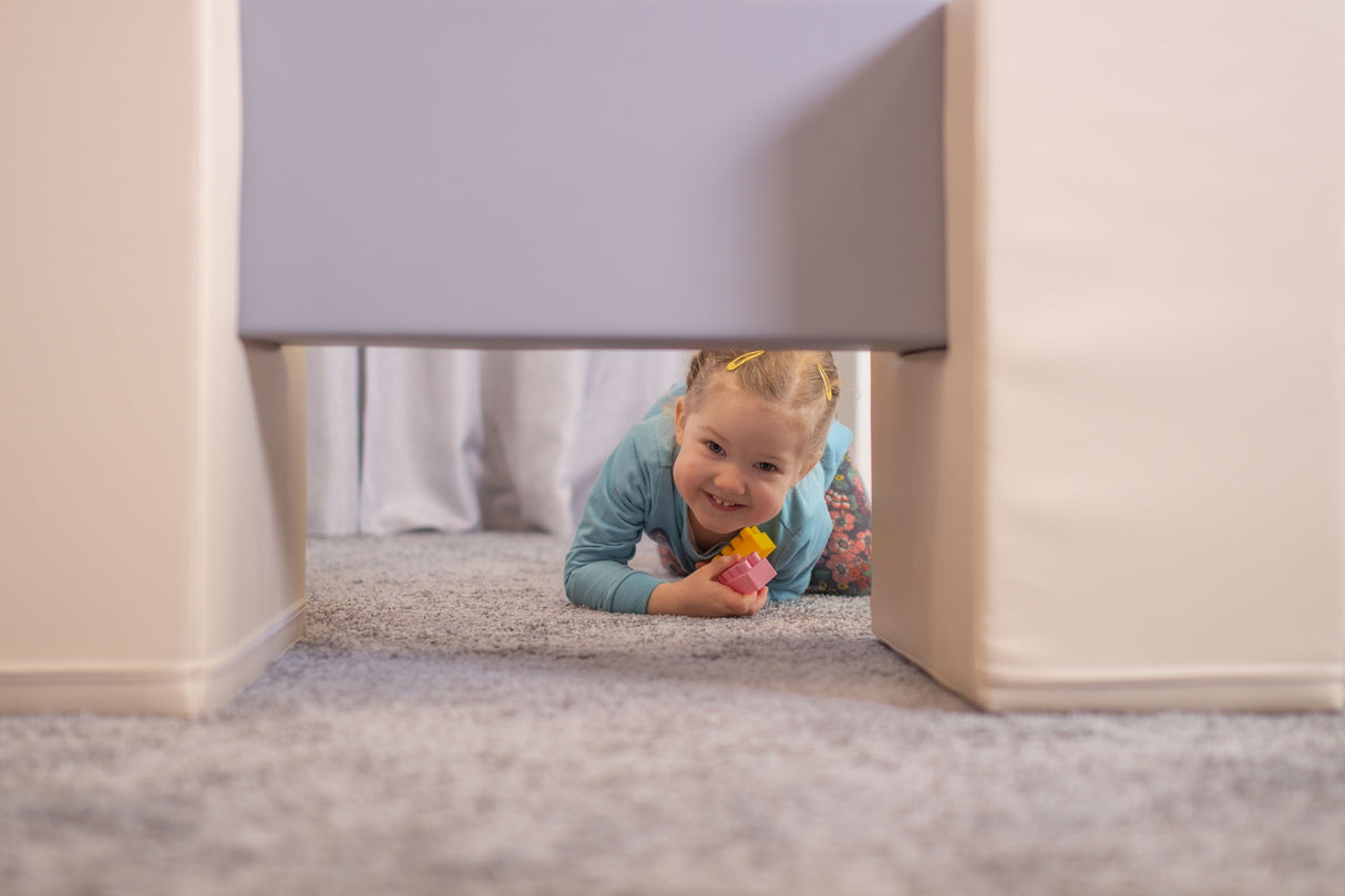 Lächelndes Kind krabbelt unter einem Möbelstück hindurch auf Teppichboden