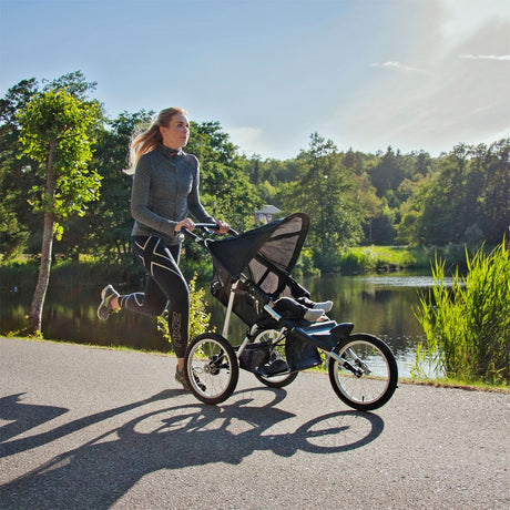 Frau joggt bei Sonnenschein mit einem dreirädrigen Jogger-Kinderwagen am See entlang
