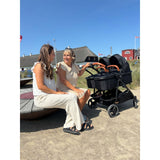 Zwei Frauen sitzen neben einem schwarzen Kinderwagen auf einem Platz